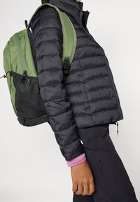 Sort pufferjakke båret over en lyserød langærmet skjorte, parret med en grøn rygsæk med en netlomme og lynlåse.