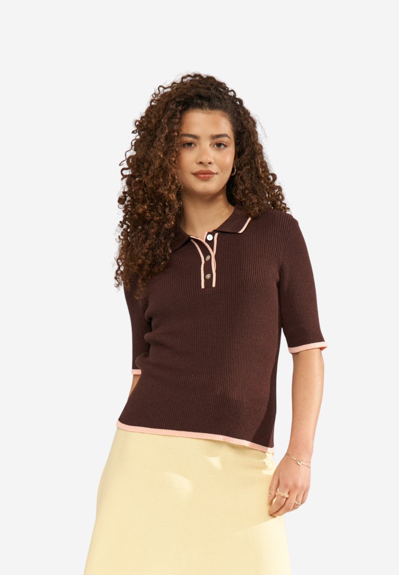 Femme aux cheveux bouclés portant un polo en maille marron à manches courtes avec un bord rose clair et une jupe jaune pâle, debout avec une main détendue.