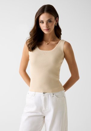Jeune femme aux longs cheveux bruns portant un haut beige sans manches en côte et un pantalon blanc, debout devant un fond clair uni.