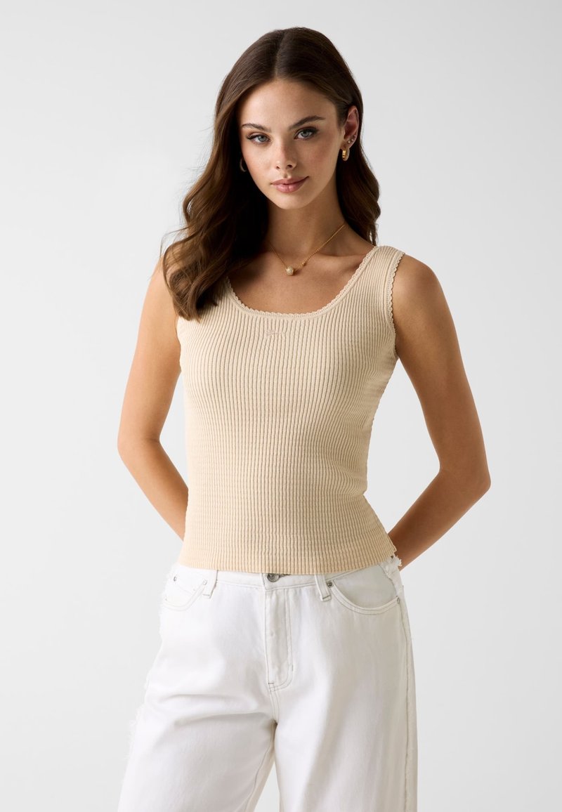 Jeune femme aux longs cheveux bruns portant un haut beige sans manches en côte et un pantalon blanc, debout devant un fond clair uni.
