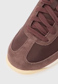 Κλείσιμο ενός δακτύλου καφέ σουέντ και υφασμάτινου αθλητικού παπουτσιού με κορδόνια που ταιριάζουν και μπεζ λαστιχένια σόλα πάνω σε ανοιχτό φόντο.