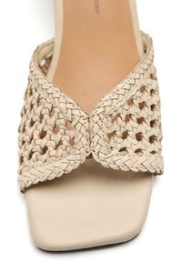 Sandalia deslizante de tejido beige con forma de punta cuadrada, que presenta detalles trenzados texturizados a lo largo del empeine y una suela plana y lisa.