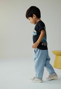 Poika, jolla on päällään musta t-paita, jossa on värikäs graafinen kuvio, vaaleansiniset farkut ja beigekengät, seisoo keltaisen jakkaran vieressä.