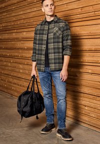 Camisa de franela a cuadros en verde y negro, camiseta negra, jeans azules, bolsa de mano negra y botas marrones con suelas blancas, todo contra una pared de madera.