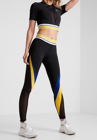 Kvinna i svarta högmidjade leggings med gula och blå diagonala ränder, en svart croppad topp med matchande ränder och vita sneakers.