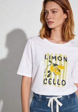 Junge Frau mit schulterlangem roten Haar trägt ein weißes T-Shirt mit Zitronengrafik und dem Text "LIMON CELLO", kombiniert mit blauen Jeans.