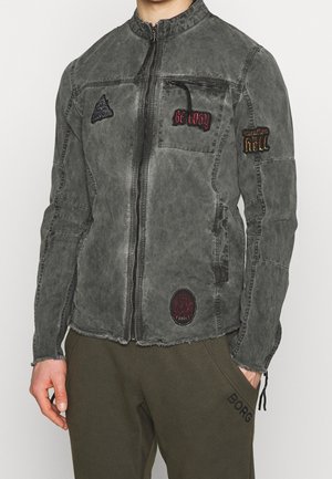Homme portant une veste grise délavée zippée avec des patches cousus et un pantalon vert foncé avec "BORG" imprimé près de la poche.