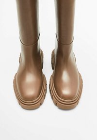 Bruine kniehoge laarzen met stevige, grof profielzolen, een gladde leren afwerking en afgeronde neuzen, weergegeven vanuit een bovenaanzicht van voren.