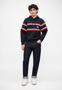 Sudadera marinera con rayas rojas y blancas, con un parche de logo en el frente. Combinada con jeans de mezclilla oscuros y zapatillas blancas.