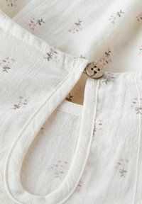 Tessuto bianco con delicata stampa floreale in rosa e verde. Presenta un bottone, un colletto curvo e dettagli di cucitura visibili.
