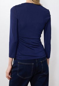 Vrouw in een getailleerde marineblauwe longsleeve top en donkerblauwe jeans, gezien van achteren tegen een effen achtergrond.