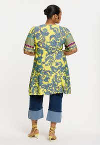 Gelbe Tunika mit blauen und grünen Paisley-Mustern, kombiniert mit lässigen, knöchelfreien Denim-Hosen und gelben Riemchen-High-Heels, von hinten betrachtet.