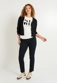 Cardigan noir sur un t-shirt blanc avec imprimé "PARIS MOOD", associé à un pantalon slim-fit bleu marine et des chaussures de sport beige/noir.