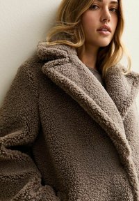 Ung kvinna som bär en tjock, texturerad brun lammskinnsjacka, ser något åt sidan mot en neutral ljus bakgrund.