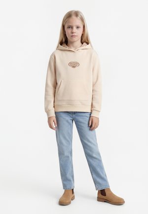 Barn med langt blond hår iført beige hettegenser, lyseblå jeans og brune ankelstøvler, stående mot en ensfarget hvit bakgrunn.