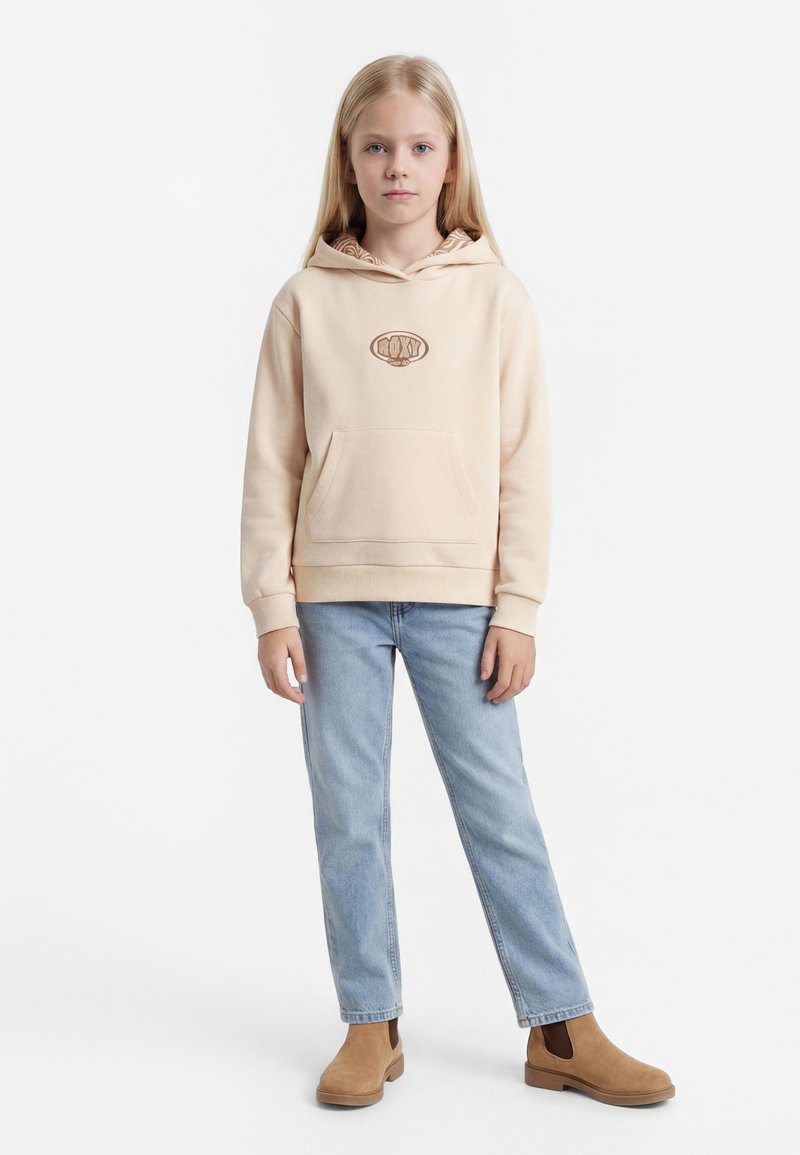 Laps pikkade blondide juustega, kes kannab beeži kapuutsiga pusa, helesiniseid teksaseid ja pruunikaid pahkluu saapaid, seisab ühevärvilise valge tausta ees.