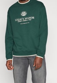 Man i mörkgrön Legacy Manor-tröja med vit text och logotyp, tillsammans med blekta grå jeans, stående mot en neutral bakgrund.