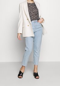 Blazer blanc croisé, haut noir à imprimé floral, jean taille haute bleu clair et mules noires à talons avec détails cloutés.