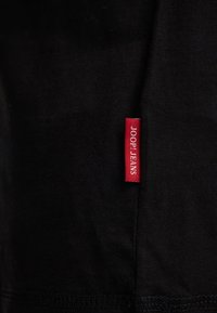 Zwart stoffen t-shirt met een opvallend etiket in het rood met de tekst "JOOP! JEANS," een gladde textuur en een eenvoudig ontwerp.