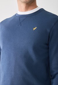 Mand iført en blå sweatshirt med rund hals og et lille gult fuglelogo på venstre bryst over en hvid t-shirt med rund hals.