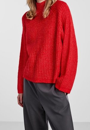 Femme portant un pull en maille rouge texturé à manches longues et un pantalon noir ample sur un fond uni.