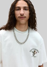 Valkoinen puuvillapaita lyhyillä hihoilla, jossa on siivekäs cherub- grafiikka ja "Guns N' Roses" -teksti. Korostettu hopealla paksulla ketjulla ja helmiketjulla.