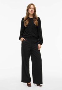Czarna teksturowana bluzka z guzikami i długimi rękawami, zestawiona z czarnymi, szerokimi spodniami z wysokim stanem oraz brązowymi szpilkami.