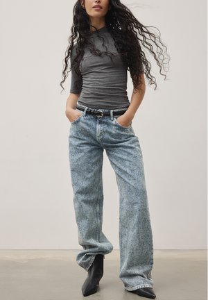 Grå kroppsnära topp med korta ärmar, ljus tvätt av vida jeans, svart bälte och svarta spetsiga ankelstövlar. Modellen står med händerna i fickorna.
