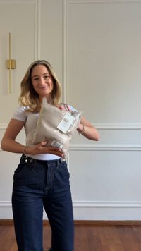 Beige stickad filt i plastförpackning, med en etikett synlig. Modell som bär en vit t-shirt och mörka denimjeans, står i ett rum.