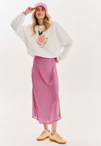 Biely sweatshirt s grafikou "Peach Please", midi sukňa s ružovými bodkami a žlté tenisky. Ružová čiapka a viacnásobné náhrdelníky dotvárajú vzhľad.