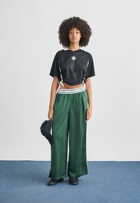 Μαύρο cropped τοπ από δίχτυ με κοντά μανίκια, συνδυασμένο με πράσινο σατέν παντελόνι φαρδιάς γραμμής με λευκή ελαστική μέση. Μαύρα επίπεδα παπούτσια.