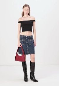 Top cropped nero off-shoulder, gonna A-line in denim con bottoni frontali e tasche, borsa rossa, stivali neri lucidi fino al ginocchio.