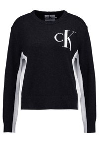 Svart och vit stickad tröja med rund halsringning, kontrasterande vita sidopaneler och 'CK'-logotyp i vitt på framsidan. Texturerade ribbade ärmslut.