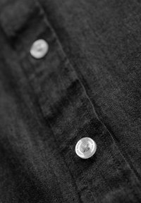 Gros plan sur un tissu noir avec deux boutons blancs cousus sur une couture verticale, le bouton inférieur gravé avec "ombre wear."