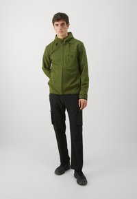 Haut à fermeture éclair vert avec une finition texturée, comportant une poche avant et une capuche ajustable. Associé à un pantalon cargo noir et des chaussures foncées.