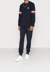 Marineblaue Langarmbluse mit rot-weißen Streifen an den Ärmeln, kombiniert mit marineblauen Jogginghosen mit einem dezenten Logo. Weiße Sneakers runden den Look ab.