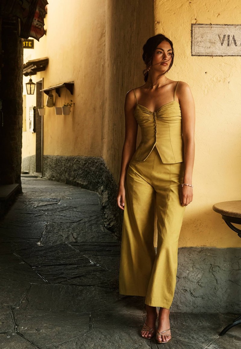Femme en haut sans manches jaune et pantalon large, debout à un coin de rue pavé, à côté d’un mur jaune avec un panneau partiel.