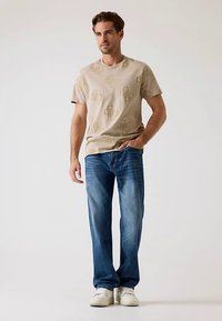 Beżowa koszulka z subtelnym wzorem, połączona z niebieskimi jeansami o prostych nogawkach. Białe trampki dopełniają stylizację. Prosty, casualowy design.