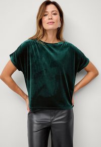 Grøn fløjlstop med en løs pasform, korte ærmer og rund halsudskæring, parret med sorte faux læderbukser. Glat tekstur og afslappet design.