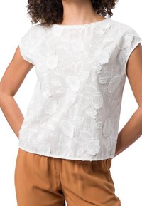 Top blanco bordado con flores, de manga corta, con cuello redondo y una tela texturada y transparente. Combinado con pantalones sueltos en tono marrón.