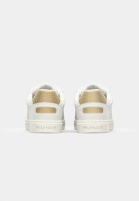 Baskets blanches avec un accent doré texturé sur le talon, présentant un détail de logo à l'arrière. Tige en cuir lisse avec une semelle plate en caoutchouc.