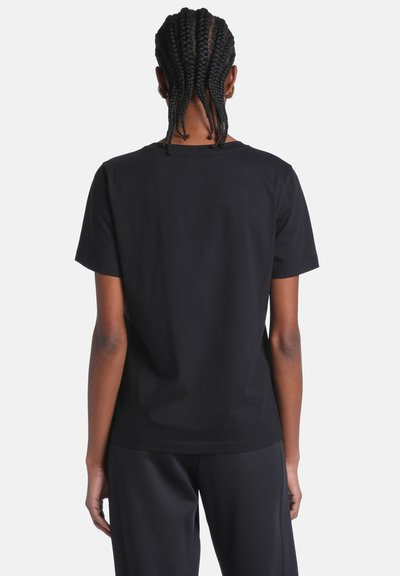 Personne aux cheveux tressés portant un t-shirt noir uni à manches courtes et un pantalon noir, vue de dos sur un fond blanc.