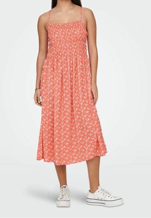 Mujer con vestido midi coral con estampado floral blanco, tirantes finos y zapatillas blancas con plataforma, de pie frente a un fondo liso.