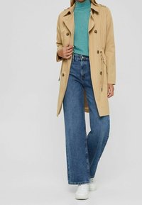 Trench-coat beige à boutons croisés, ceinture à nouer, associé à un col roulé côtelé couleur sarcelle et un jean bleu à jambes larges. Baskets blanches.