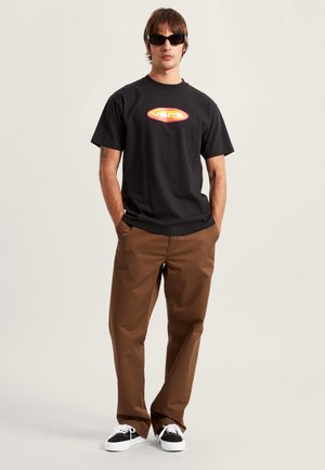 Sort bomuld T-shirt med et rødt og orange Vans-logo, kombineret med brune bukser og sorte og hvide sneakers. Modellen bærer sorte solbriller.