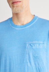 Světle modré bavlněné tričko s kulatým výstřihem, malou náprsní kapsou a vyšitým logem Adidas nad kapsou. Měkká textura.