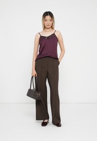 Camisole di raso viola con dettaglio di paillettes, abbinato a pantaloni larghi marroni. La modella tiene una piccola borsa scura e indossa scarpe borgogna lucide.