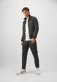 Giubbotto bomber nero con motivo, con polsini elasticizzati, indossato su una maglietta bianca, abbinato a pantaloni scuri e sneakers bianche.