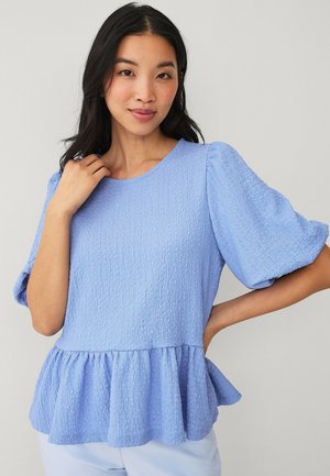 Kvinde med langt mørkt hår iført en tekstureret lyseblå bluse med pufærmer og flæsekant, poserer med den ene hånd på hoften.