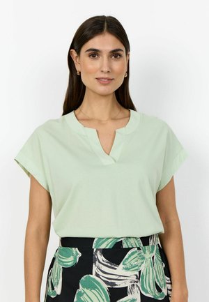 Femme aux longs cheveux bruns portant un haut vert clair à manches courtes et une jupe noire avec des motifs de feuilles vertes et blanches, debout devant un fond blanc.